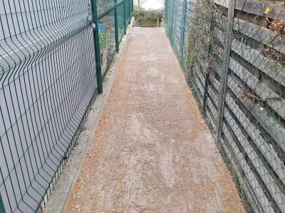 Allée étroite avec sol en gravier, clôture métallique verte sur les côtés.