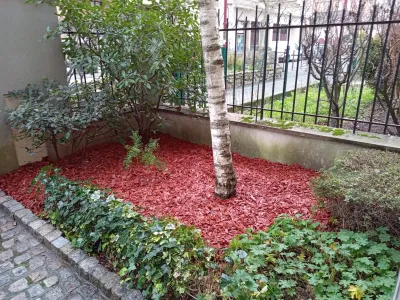 Petit massif au sol avec paillis rouge, arbre et haie derrière