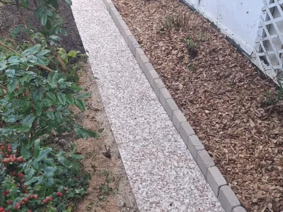 Allée piétonne en gravier clair bordée de parterres et végétation.