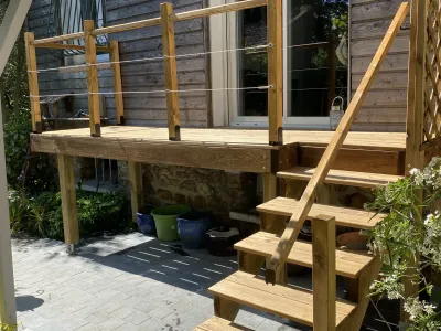 Terrasse bois surélevée avec garde-corps et escalier, maison en arrière-plan.