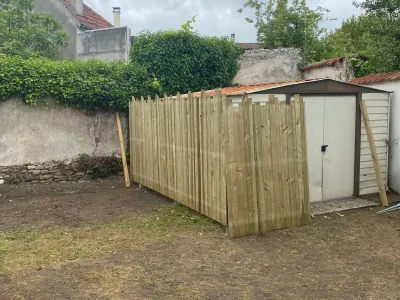 Clôture en bois récemment installée autour d’un abri de jardin, sol nu et mur en arrière-plan.