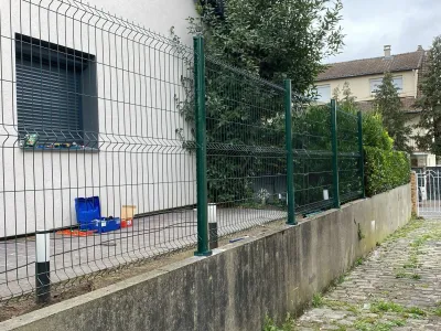 Allée longeant un bâtiment avec sol en gravier et clôture métallique verte, vue d’extérieur.